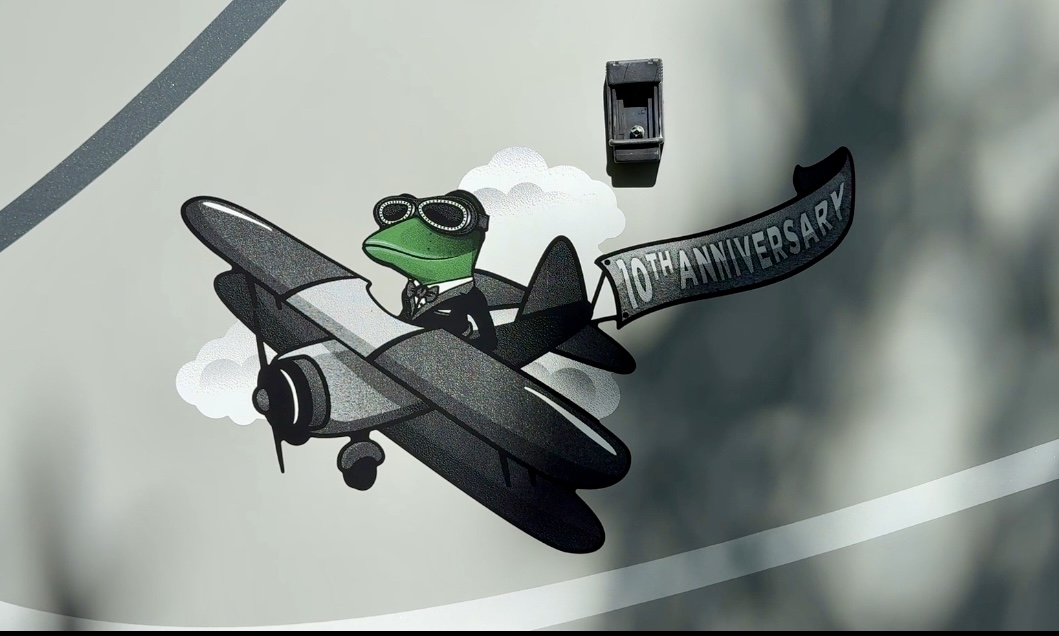 A cartoon frog in a pilot's outfit flying a small airplane, accompanied by a banner that reads '10th Anniversary'.