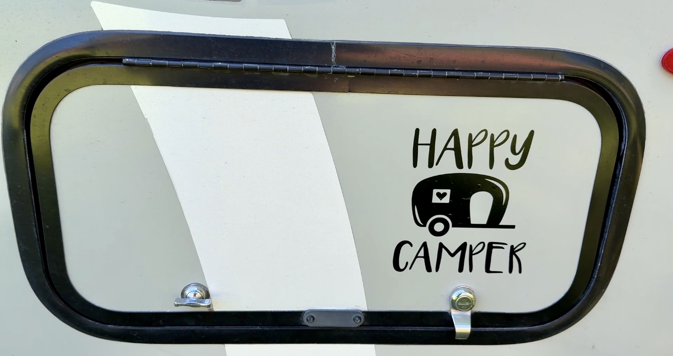 A close-up of a camper's exterior featuring a black frame window with the text 'HAPPY CAMPER' and a small graphic of a camper illustration.