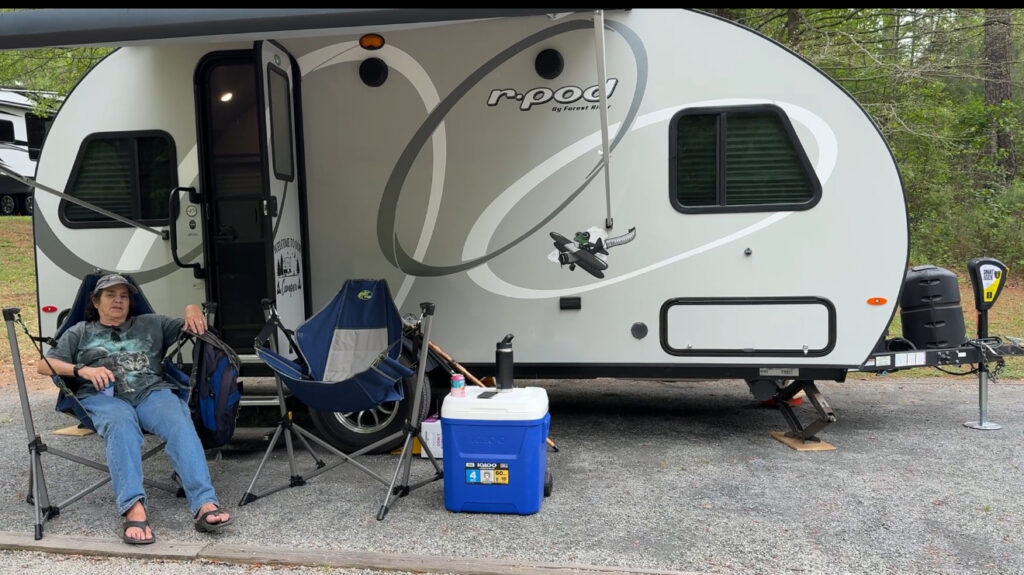 A person sitting in a camping chair next to a 2019 Forest River R-Pod camper, with a cooler and camping gear nearby.