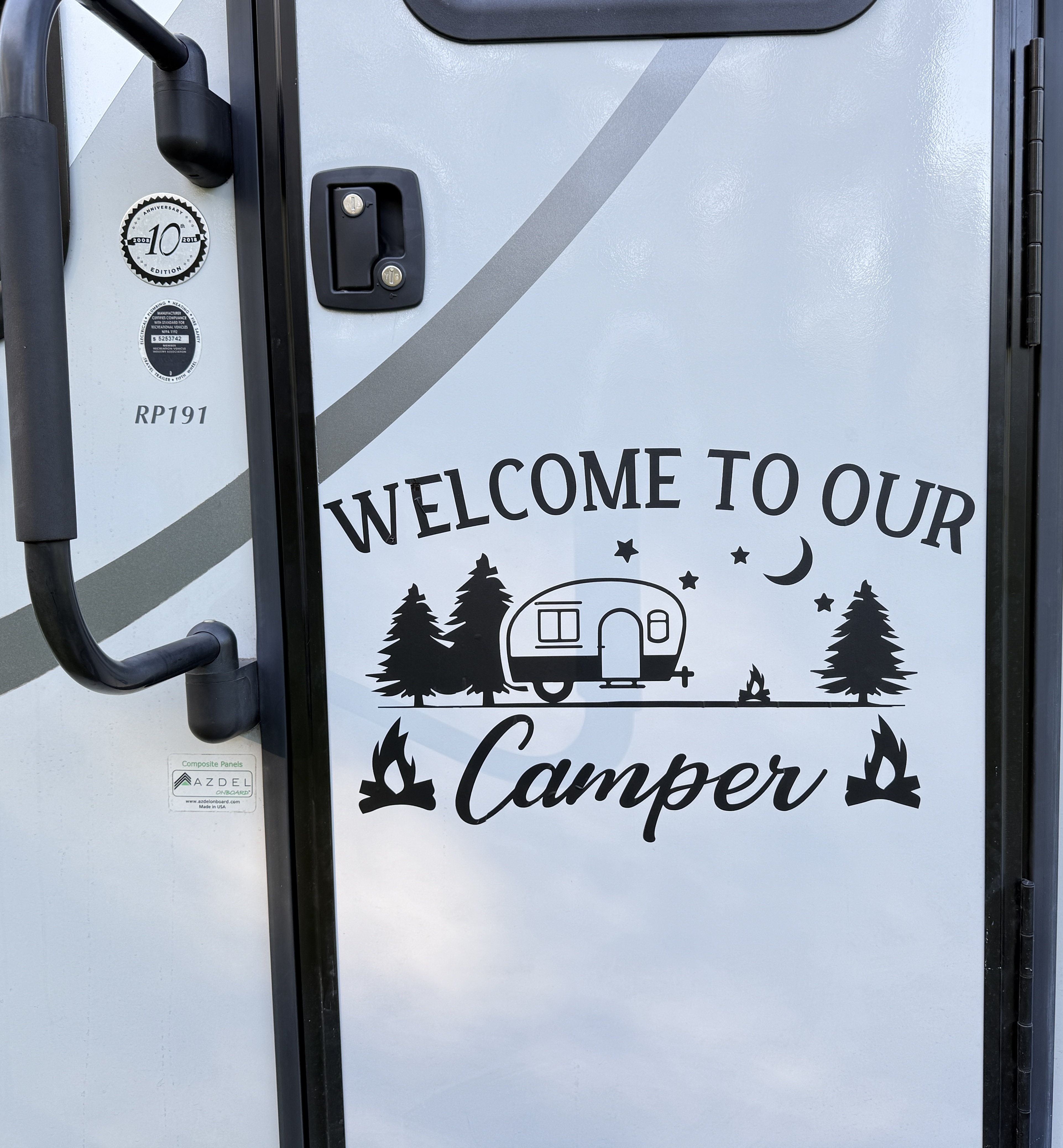 Close-up view of the door of a camper featuring a decorative sign that reads 'WELCOME TO OUR Camper,' accompanied by an illustration of a vintage trailer, trees, stars, and campfires.